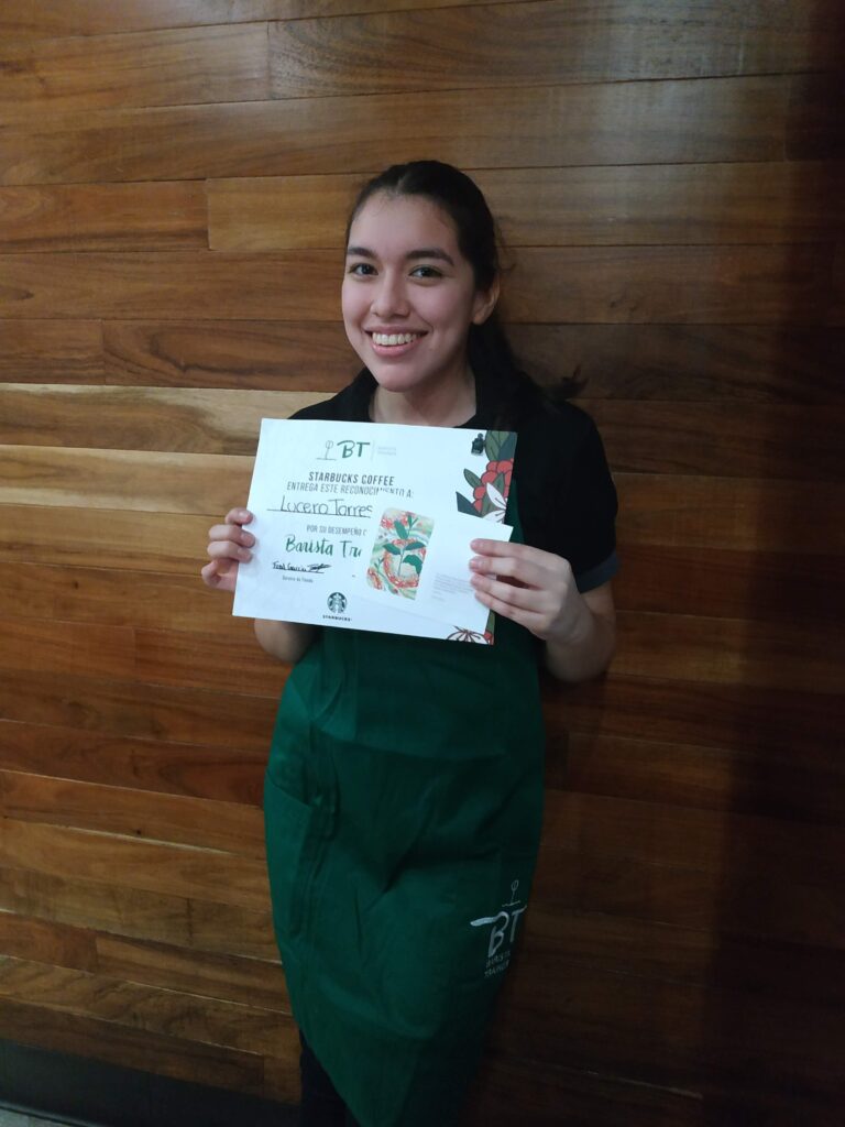 Luz smiling and holding her Starbucks Barista Trainer certificate while wearing a green apron.