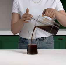 A person in a white t-shirt and jeans pours finished cold brew coffee from a glass French press into a clear carafe on a white countertop.